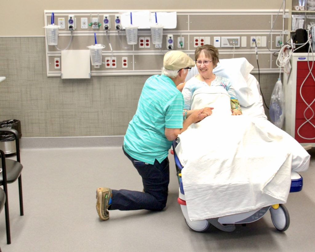 Patient in hospital bed receives care and is comforted by her husband.